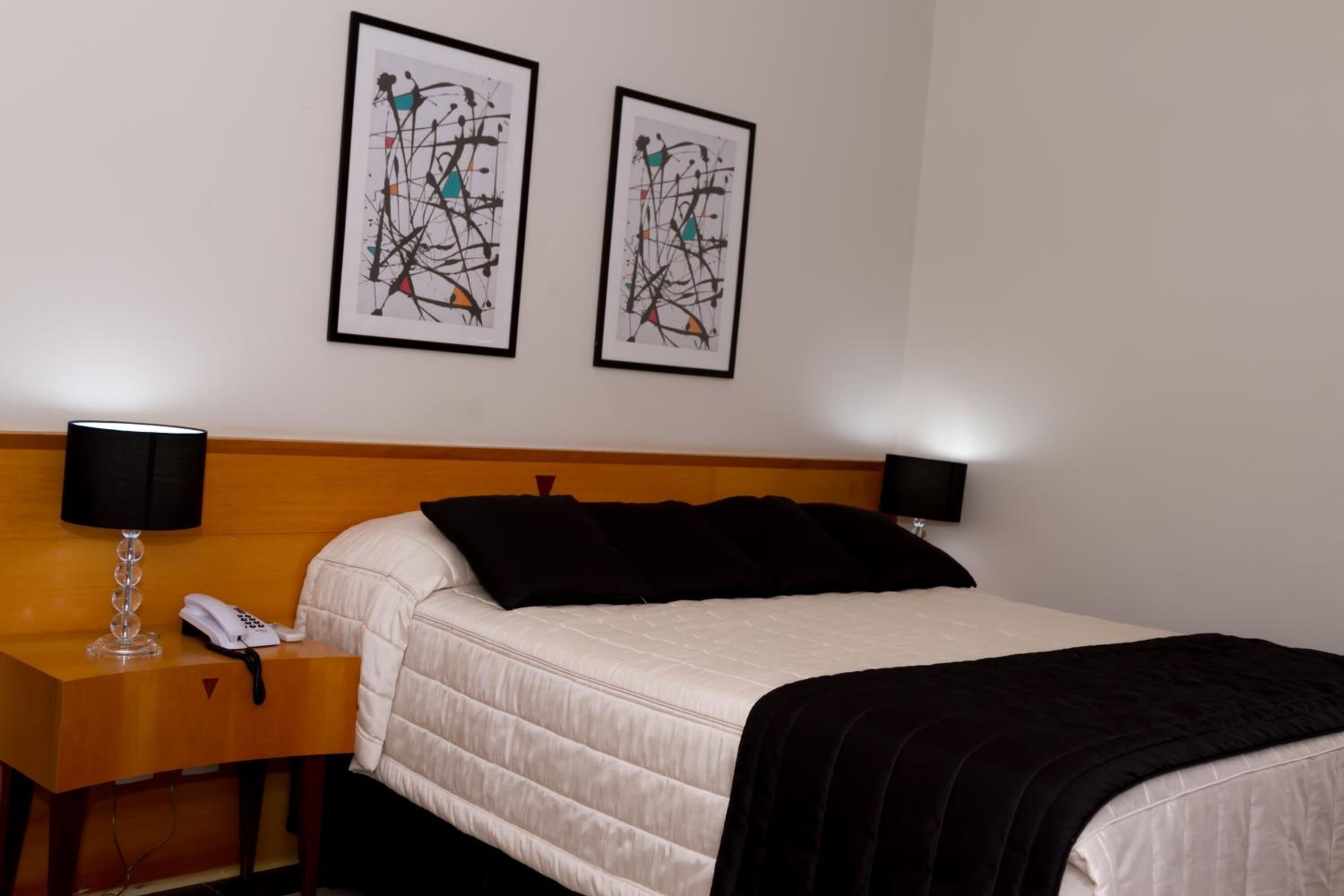 A hotel room with a bed, a bedside table with a lamp, a phone, and two abstract paintings on the wall.