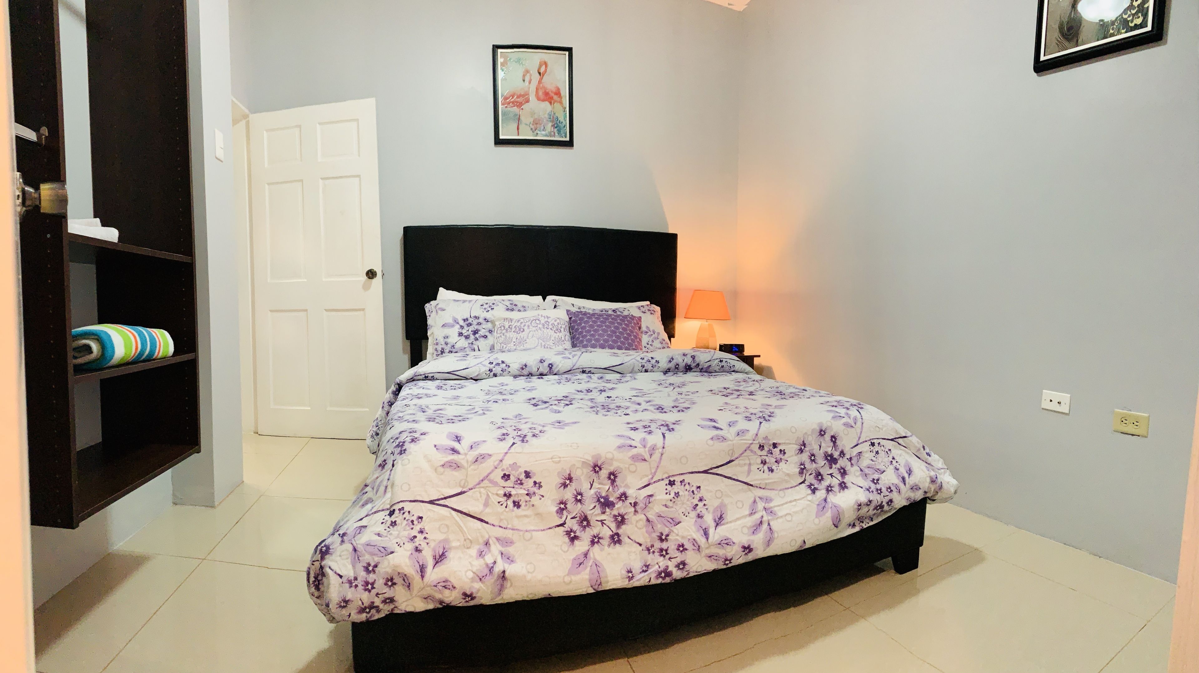 Une chambre à coucher avec un lit, une table de chevet, une lampe, une porte et une étagère avec des serviettes.