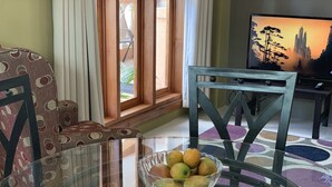 A living room with a glass coffee table, a patterned armchair, a television, and a view of the outdoors through a window. - Robbie's Place Tobago (Black Rock)