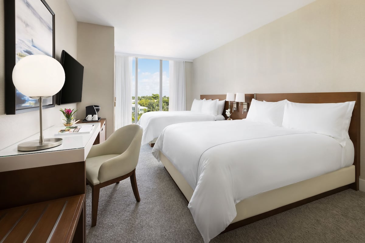 A hotel room with two beds, a desk with a chair, a flat-screen TV, and a large window offering a view of greenery.