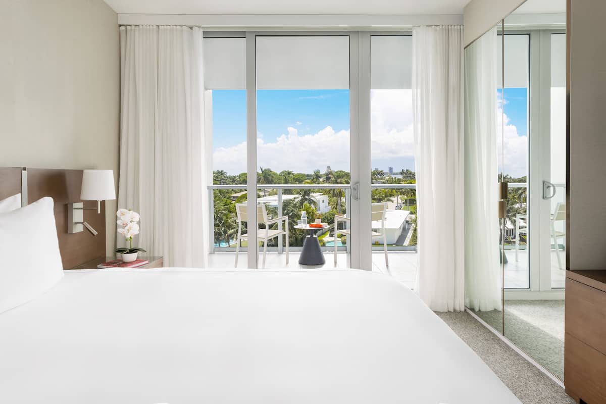 A hotel room with a large bed, a nightstand with a lamp, a potted orchid, a small table with a vase, and a balcony with a view of palm trees and buildings.
