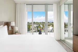 Habitación de hotel con una cama grande, una mesita de noche con una lámpara, una orquídea en maceta, una pequeña mesa con un florero y un balcón con vistas a palmeras y edificios.