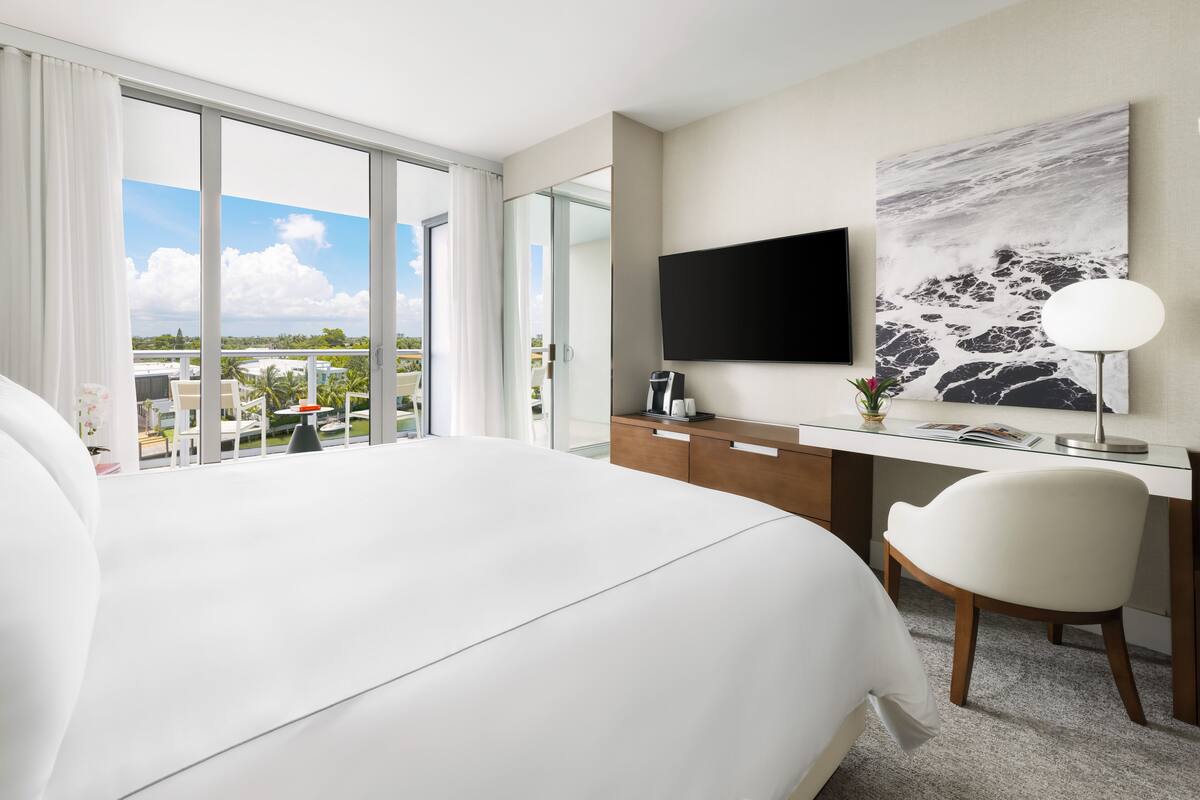 A modern hotel room with a large bed, a desk with a flat-screen TV, a chair, and a view of the city through large windows.
