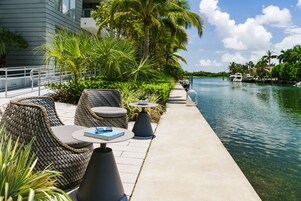 A waterfront patio with wicker furniture, a clear view of the water, and a modern building in the background.