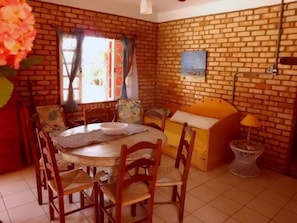 A dining area with a brick wall, a round table, wooden chairs, a bench, and a small side table with a lamp. - Recanto dos Bambus (Florianópolis)