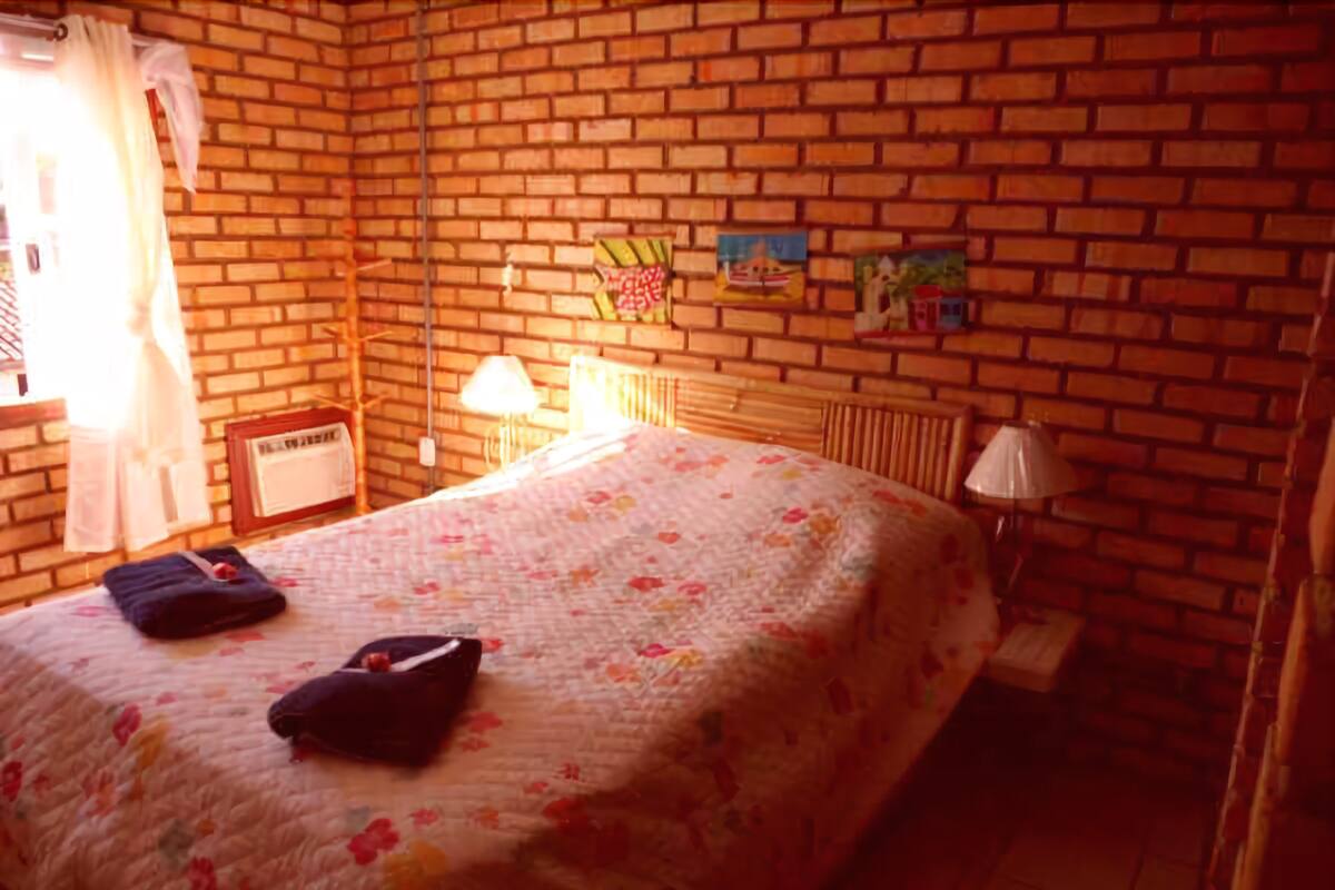Une pièce avec un mur en briques, un lit recouvert d’une couvre-lit à motifs floraux, une table de chevet, une lampe et une fenêtre avec des rideaux.