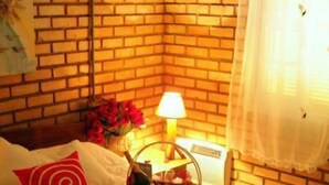 Une chambre d’hôtel avec un mur en briques, un lit recouvert de draps blancs, un oreiller rouge, une table de chevet avec une lampe et une fenêtre ornée d’un rideau léger.