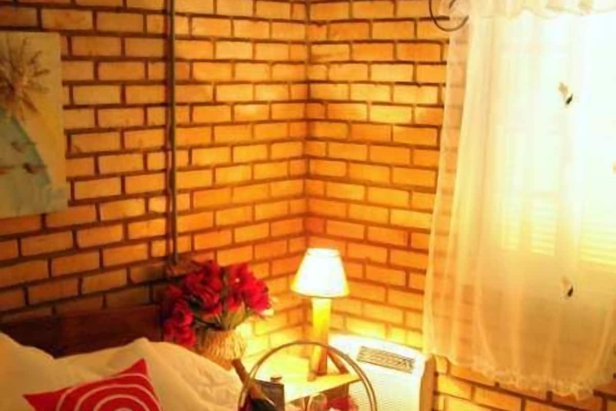 Une chambre d’hôtel avec un mur en briques, un lit recouvert de draps blancs, un oreiller rouge, une table de chevet avec une lampe et une fenêtre ornée d’un rideau léger.