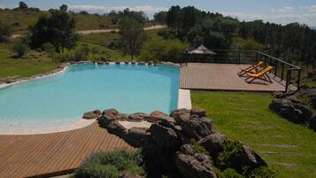 Una piscina con muelle de madera y sillones de descanso, rodeada de vegetación y colinas.