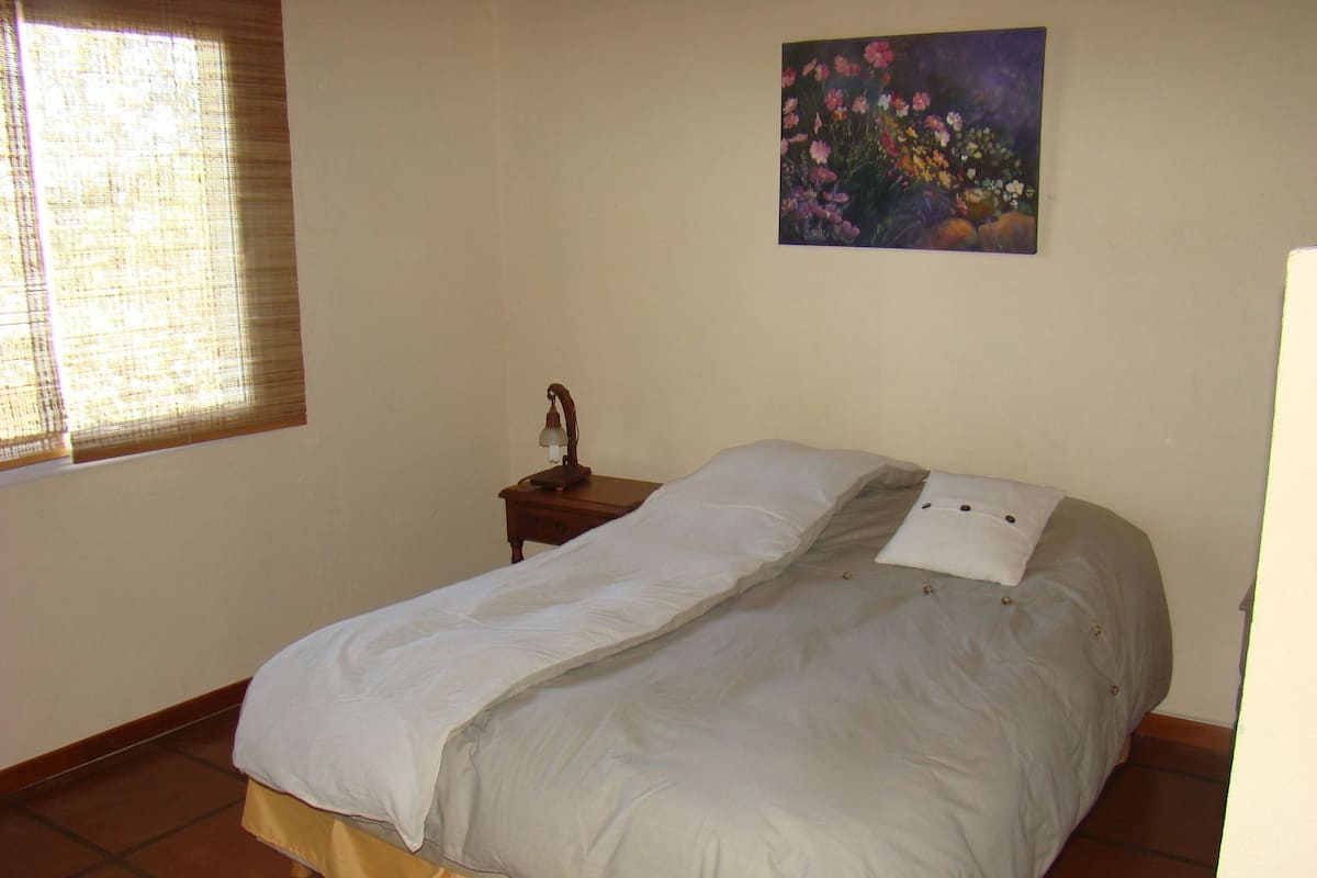 Um quarto com cama, mesa de cabeceira, janela com persianas e um quadro na parede.