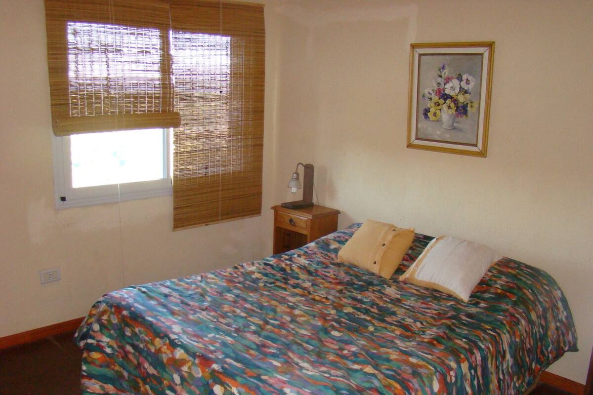 Quarto com cama, mesa de cabeceira, luminária e janela com persianas.
