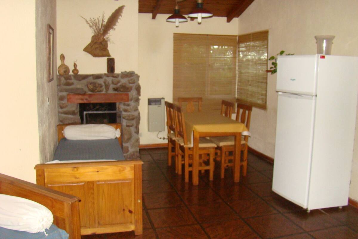 Um quarto pequeno e acolhedor com lareira de pedra, uma mesa de jantar de madeira com cadeiras, uma cama com cabeceira de madeira e uma geladeira branca.