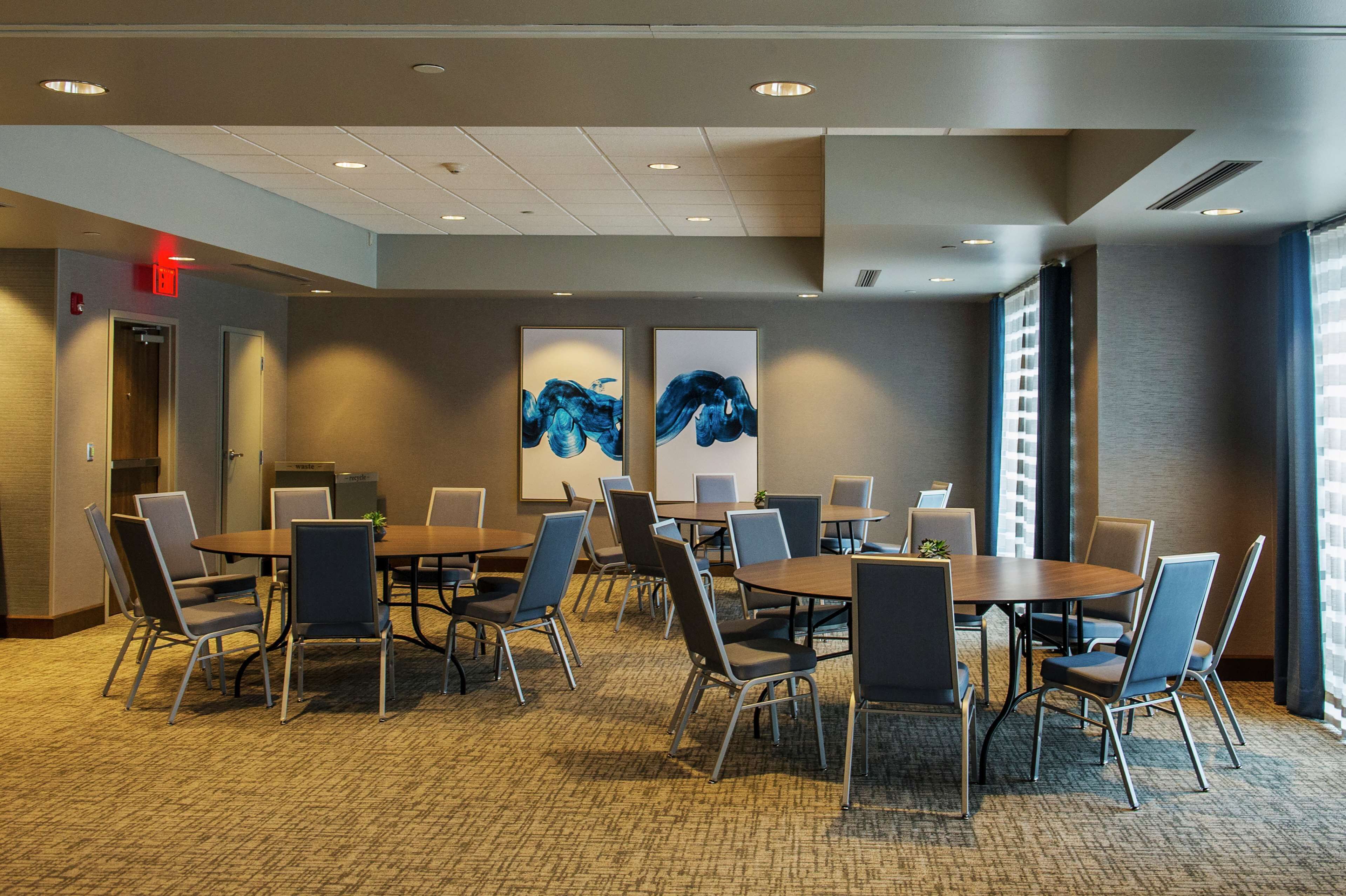 A conference room with multiple tables and chairs, large windows, and two abstract paintings on the wall.