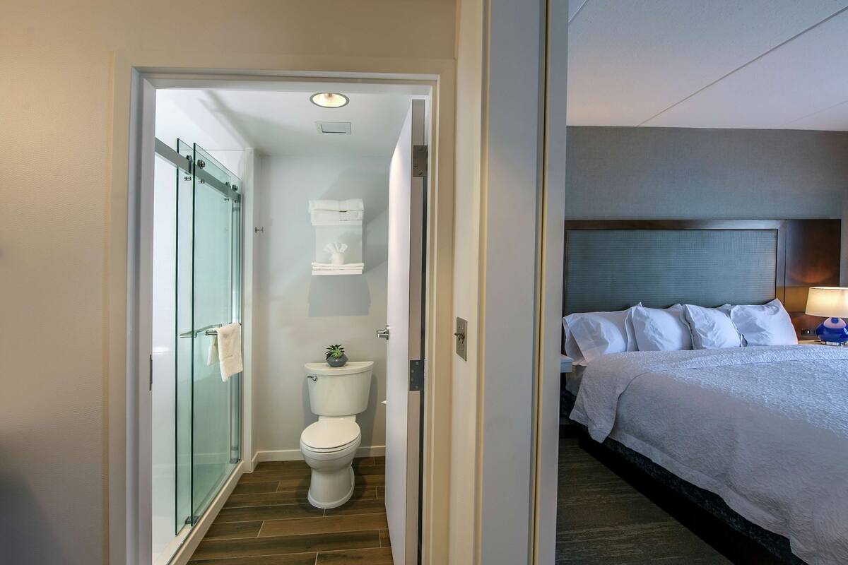 A hotel room with a glass-enclosed shower, a white toilet, and a neatly made bed with white bedding.
