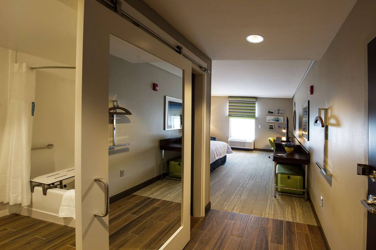 A hotel room with a shower, a bed, a desk with a chair, and a window with blinds.