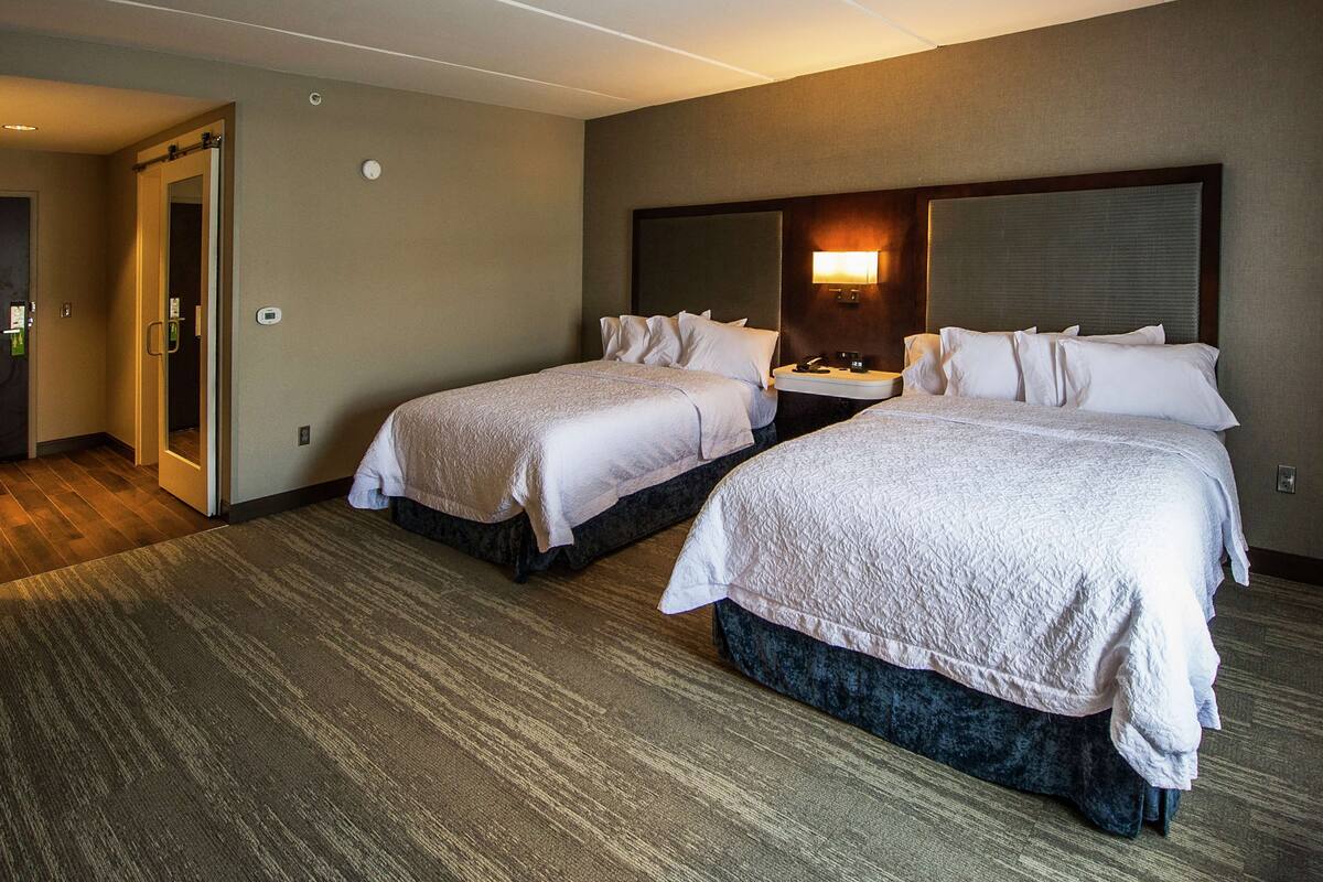 A hotel room with two beds, a bedside table with a lamp, and a door leading to another room.