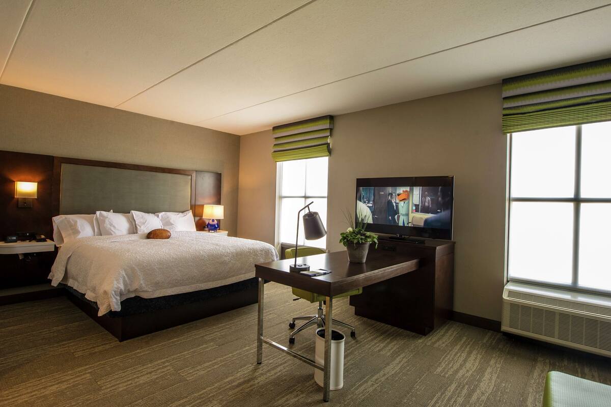 A hotel room with a bed, desk, television, lamp, and windows.