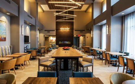 A spacious dining area with multiple tables and chairs, a central wooden table with a fruit bowl, and modern lighting fixtures.
