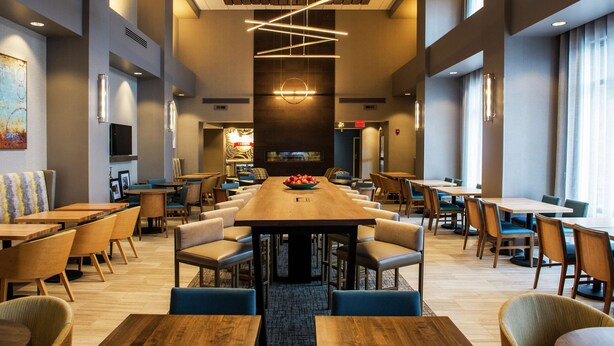 A spacious dining area with multiple tables and chairs, a central wooden table with a fruit bowl, and modern lighting fixtures. - Hampton Inn & Suites Foxborough/Mansfield (Foxborough)