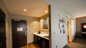 A modern hotel hallway with a wooden floor, a dark door, a white sink with a mirror, and framed pictures on the wall. - Hampton Inn & Suites Foxborough/Mansfield (Foxborough)