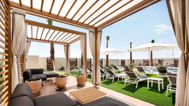 A covered outdoor seating area with lounge chairs and umbrellas, a pool in the background, and potted plants. - Embassy Suites by Hilton San Antonio Landmark (San Antonio)