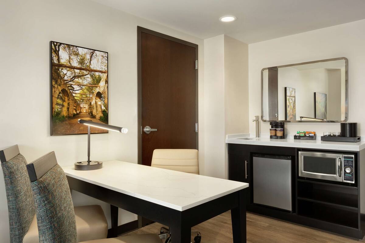 A modern kitchen with a microwave, coffee maker, and a refrigerator. There is a dining table with a lamp and a chair. A door with a mirror and artwork is visible.