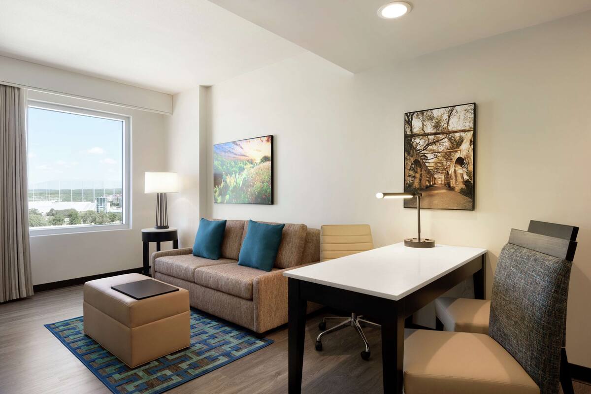 A hotel room with a sofa, ottoman, table, and chairs. There is a window with curtains, a lamp, and artwork on the wall.