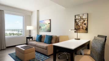 Quarto de hotel com sofá, banquinho, mesa e cadeiras. Possui janela com cortinas, luminária e uma obra de arte na parede.
