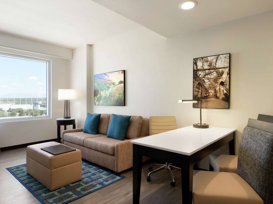 Quarto de hotel com sofá, banquinho, mesa e cadeiras. Possui janela com cortinas, luminária e uma obra de arte na parede.