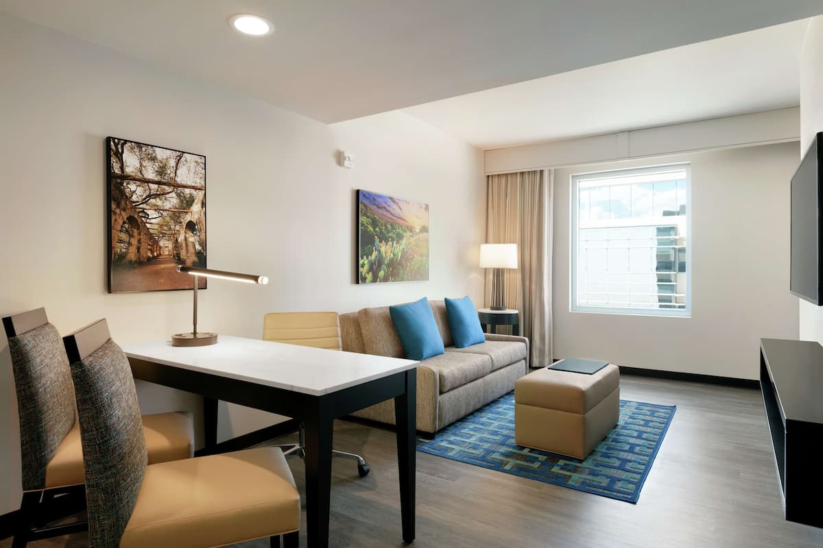 A modern hotel room with a dining table, sofa, and ottoman, a TV, and artwork on the walls.