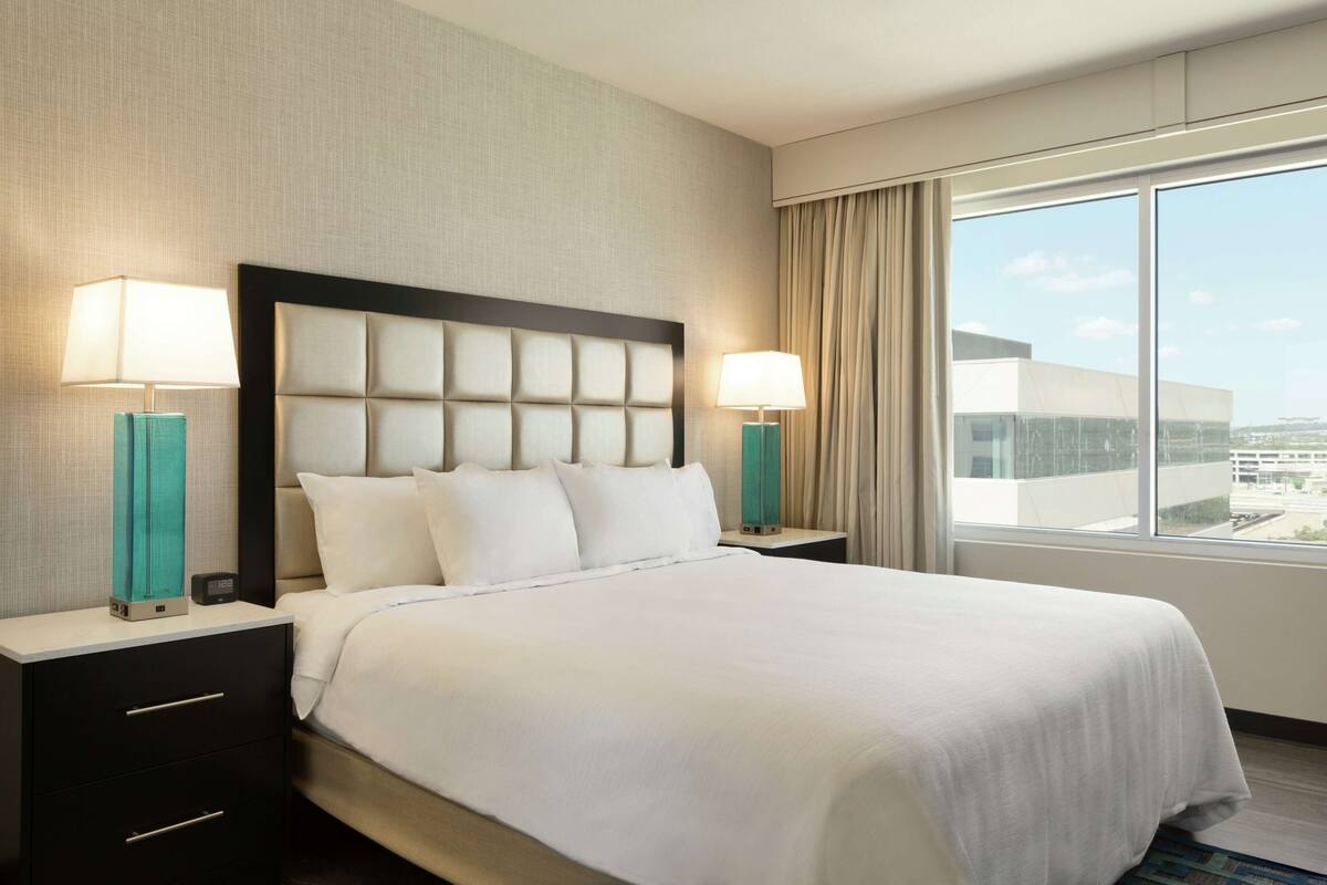 A hotel room with a large bed, two bedside tables with lamps, a view of the city through the window, and a modern architectural style.