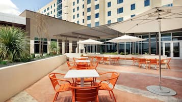 An outdoor patio area with orange chairs and white tables, surrounded by a modern building with large windows.