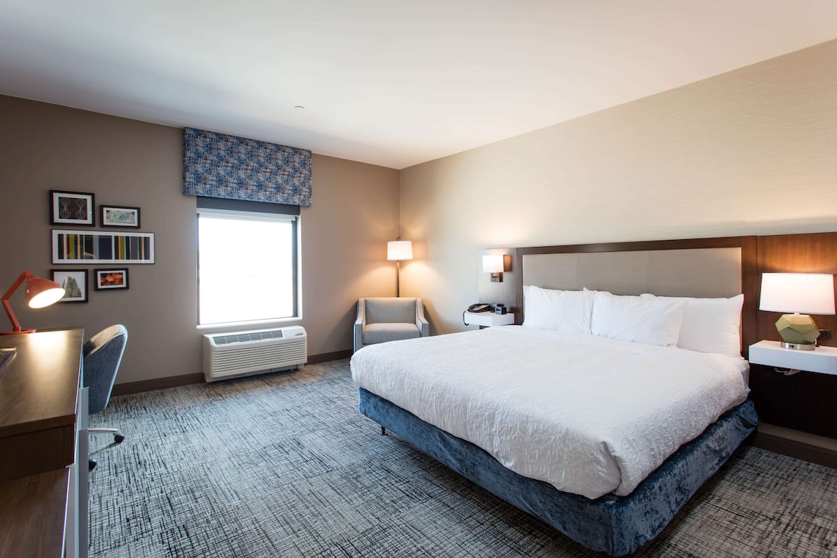 A hotel room with a large bed, a desk, a chair, a window with blinds, a wall-mounted lamp, and a wall-mounted air conditioner.