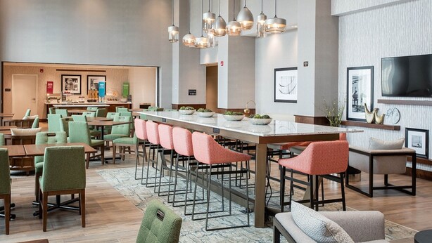 A modern hotel lobby with a long communal dining table, multiple seating areas, and a large flat-screen TV mounted on the wall. - Hampton Inn & Suites St. George, UT (St. George)