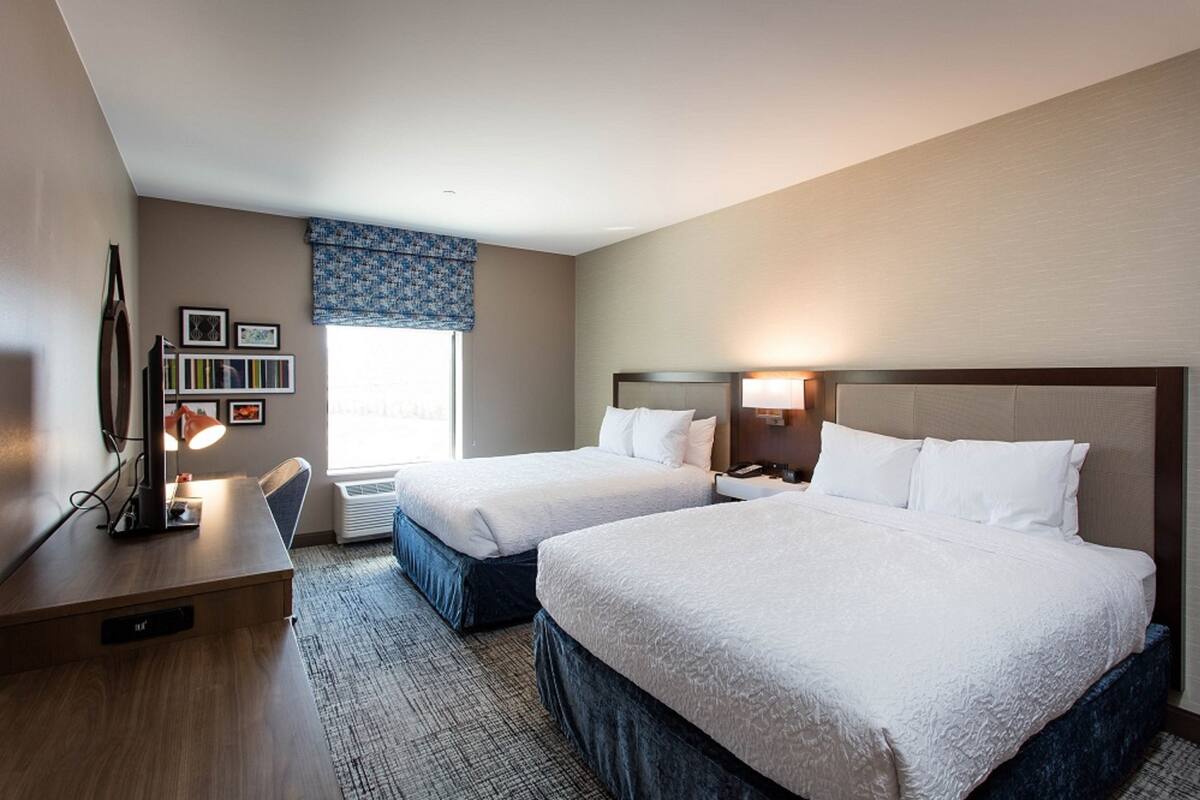 A hotel room with two beds, a desk with a lamp, a bookshelf, and a window with a view.