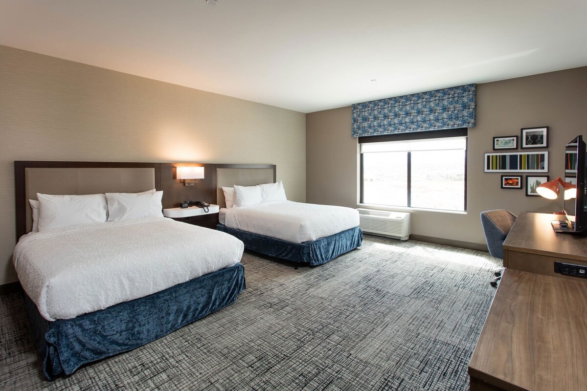 A hotel room with two beds, a desk, a chair, a window with a view, and a wall with framed pictures.