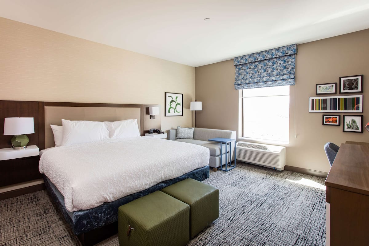 A hotel room with a large bed, a desk, a chair, a window with a view, and a wall with framed pictures.