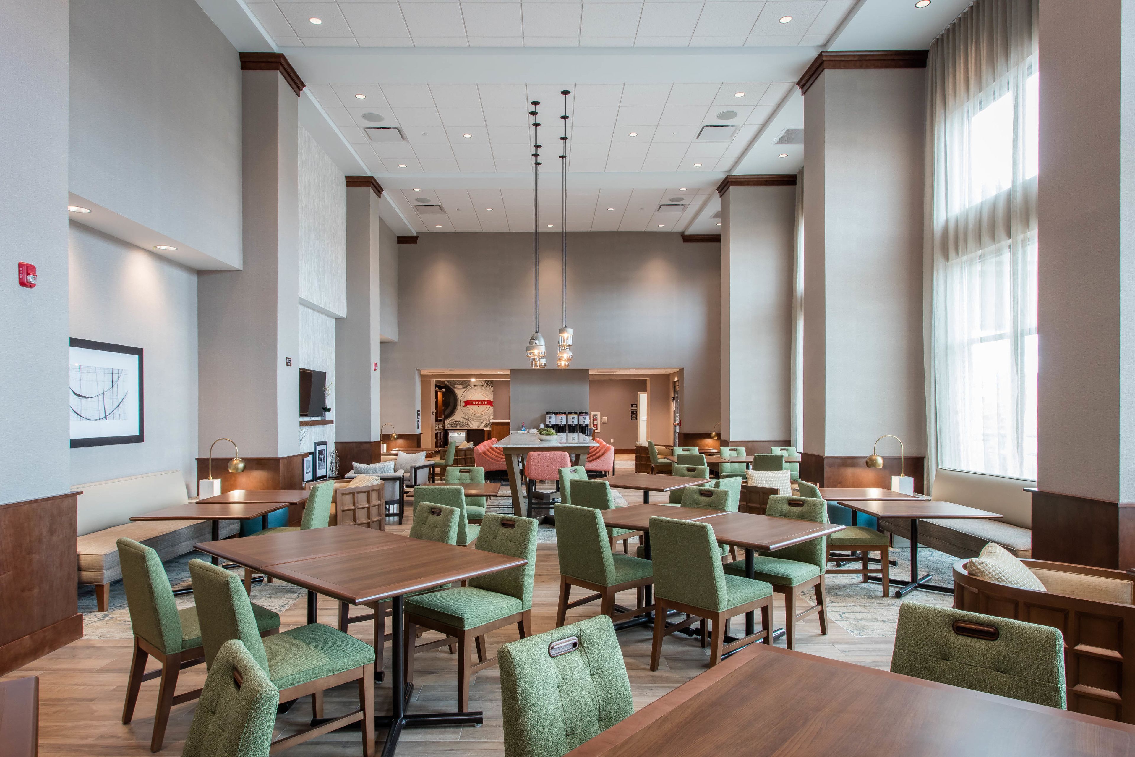 Un vaste espace de restauration dans l’hôtel, avec de nombreuses tables et chaises, un bar central et de grandes fenêtres laissant entrer la lumière naturelle.