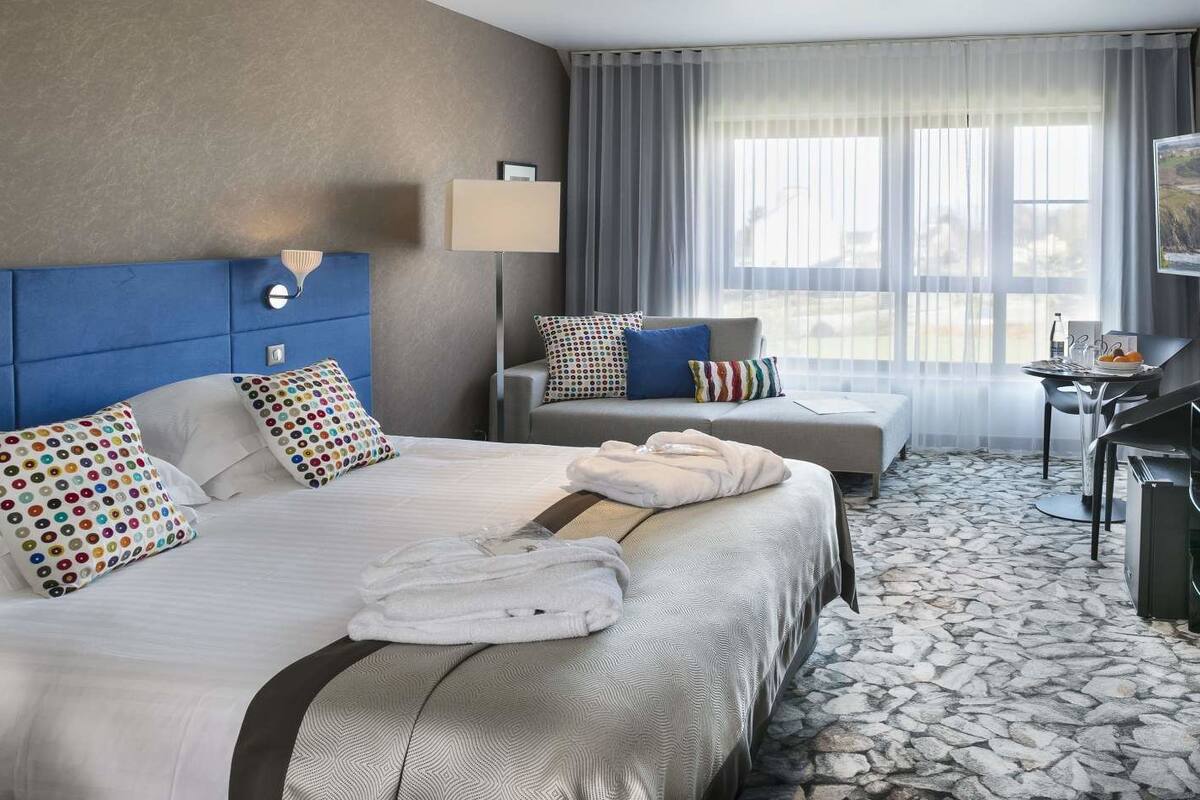 Ein Hotelzimmer mit zwei Betten, einem Sofa, einem Schreibtisch und einem großen Fenster mit Vorhängen.