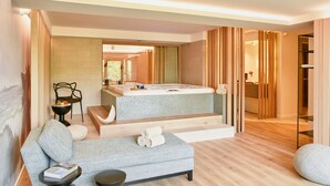 Quarto de hotel moderno com uma cama grande, área de estar com sofá e banquinho, uma mesinha e uma janela ampla.