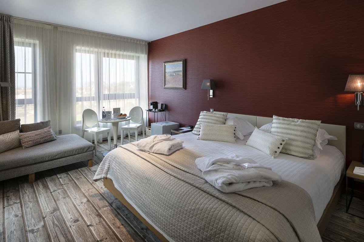 Ein Hotelzimmer mit einem großen Bett, einer Sitzecke, einem kleinen Tisch und einem Sessel. Ein Fenster mit Vorhängen und ein Gemälde an der Wand sind ebenfalls vorhanden.