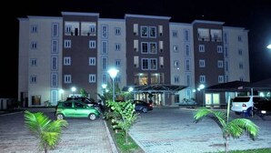 A multi-story hotel with a central entrance, a parking area with cars, and small palm trees. - De Edge Hotel (Port Harcourt)