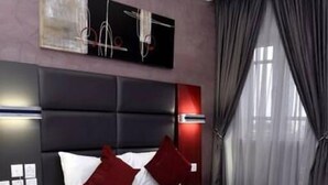 A modern hotel room with a large bed, red and white bedding, a dark headboard, a bedside table with a lamp, and a view of the city through large windows. - De Edge Hotel (Port Harcourt)