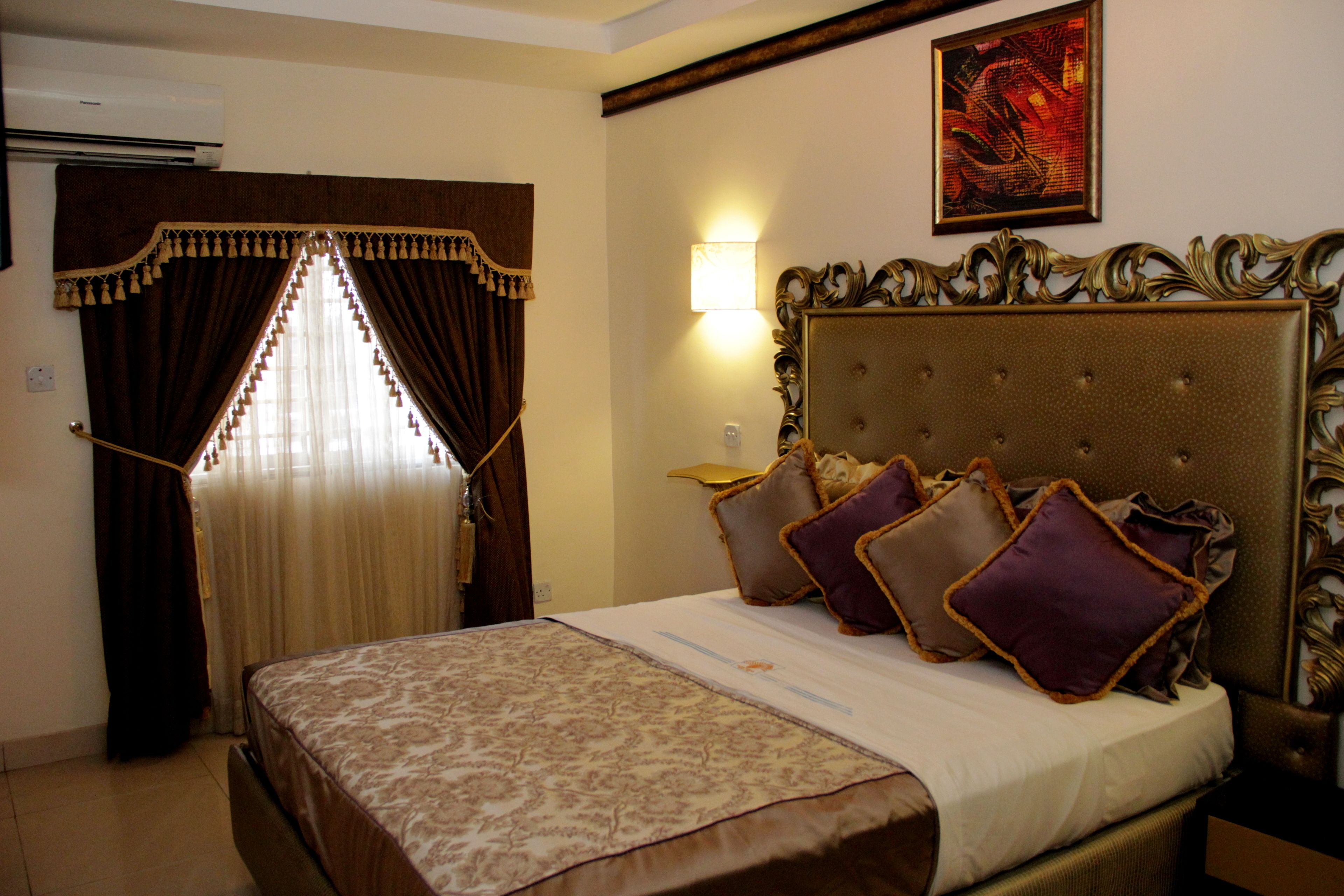 Une chambre d’hôtel avec un grand lit, une tête de lit ornée de motifs décoratifs, une lampe fixée au mur et un tableau au-dessus du lit.