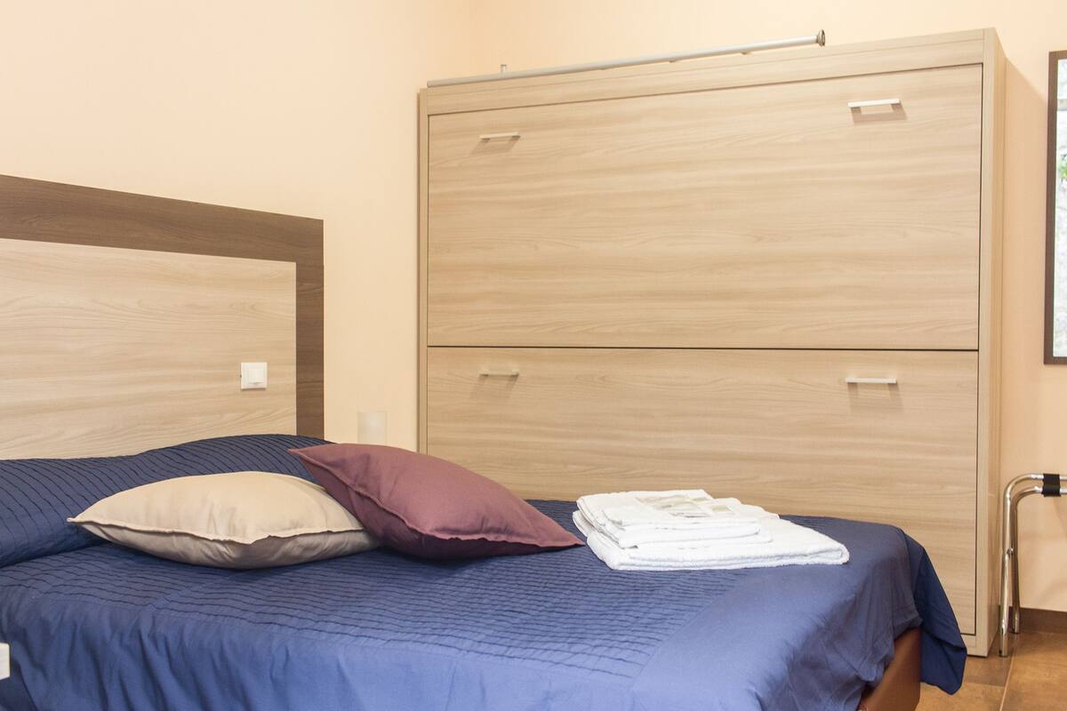 Una cama con una colcha azul, un almohadón color beige y un almohadón morado. Se observa un armario de madera con dos puertas y un tirador metálico.