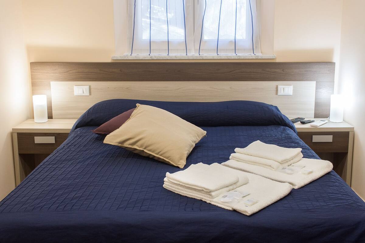 Una cama bien hecha con colcha azul, almohada color beige y almohada marrón. Hay toallas dobladas sobre la cama. La habitación tiene cabeceras de madera, lámparas de noche y una ventana con persianas.