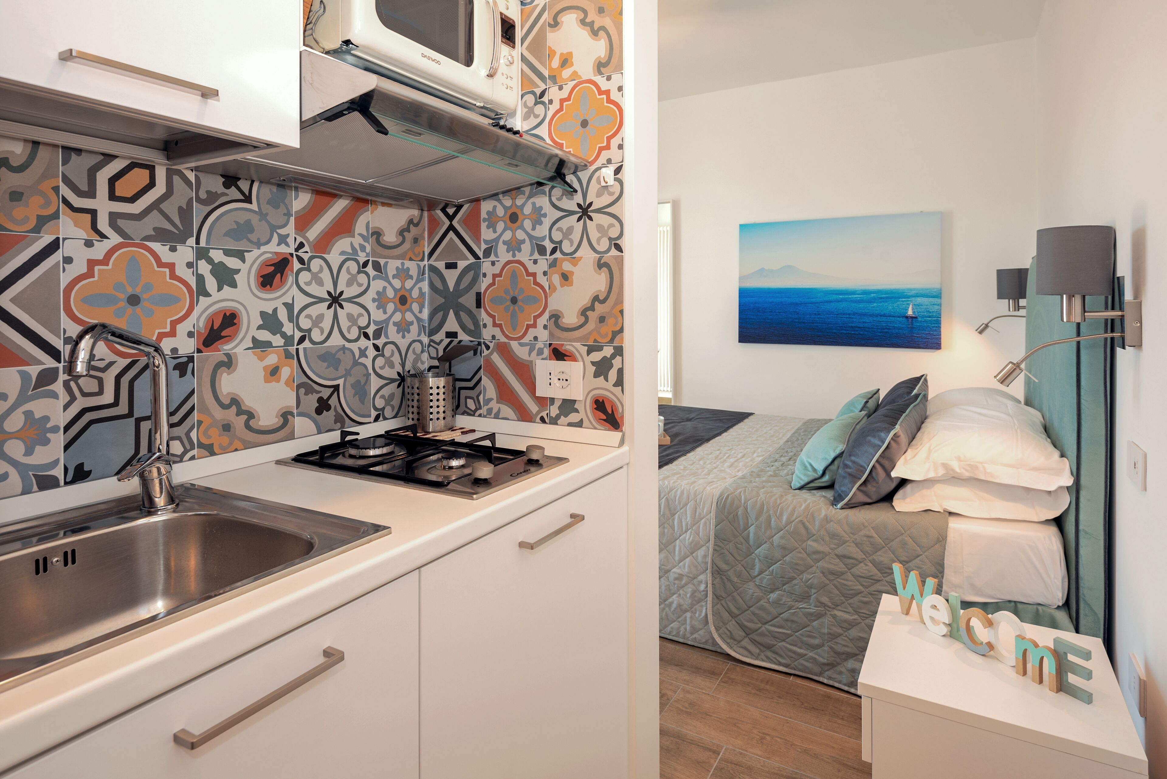A compact kitchen with a modern stove, a stainless steel sink, and a microwave. The room features a bed with a quilted cover and a painting of a seascape.