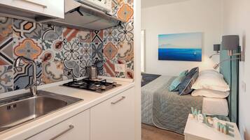 A compact kitchen with a modern stove, a stainless steel sink, and a microwave. The room features a bed with a quilted cover and a painting of a seascape.