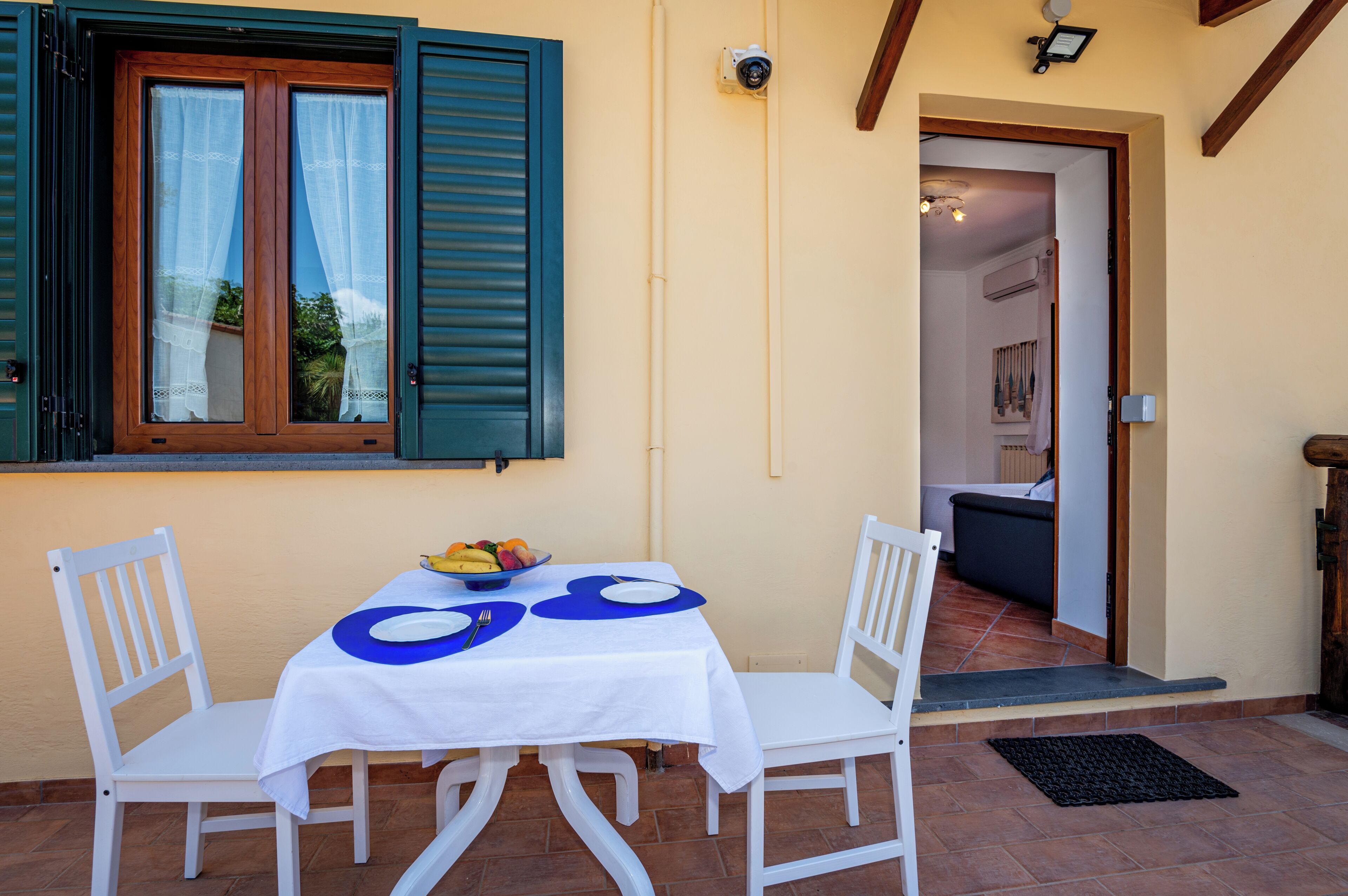 A dining area with a table set for two, a bowl of fruit, and a view of the interior through an open door.