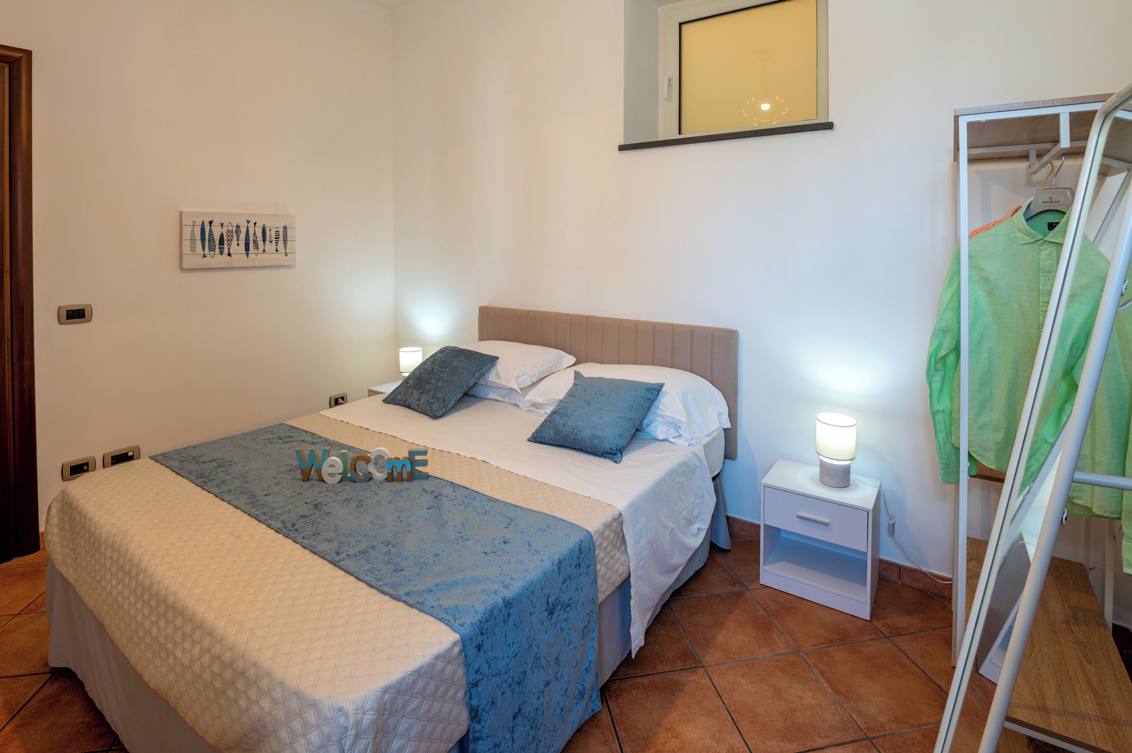Un lit bien fait, recouvert d’une couvre-lit bleue et blanche, une table de chevet avec une lampe et un miroir sur lequel est accrochée une serviette verte.